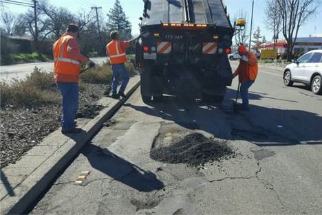 Road maintenance workers in orange safety vests repairing a large pothole beside a dump truck, spreading the asphalt on a damaged section of pavement.