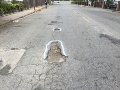 A cracked asphalt road with several large potholes outlined in white paint, extending down the middle of the street.